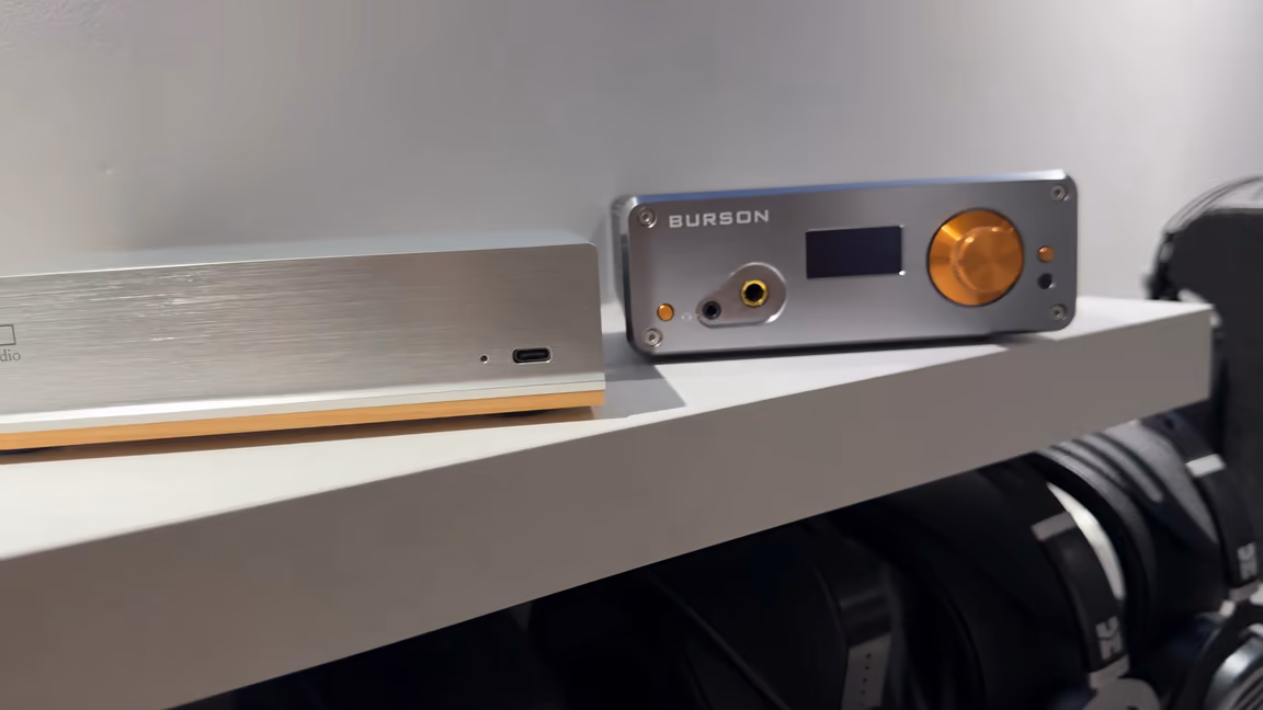 The Henry Audio DA 256 on a desk shelf next to a Burson headphone amplifier, with headphones hanging below, showing how low and shallow the DAC is beside a normal desktop amp