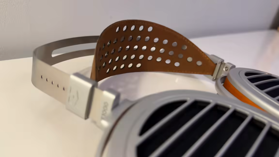 The HIFIMAN HE1000 Stealth's suspension headband: the CNC-machined aluminium band and slider marked HE1000, holding the perforated tan leather strap, with a wood-accented ear cup at right