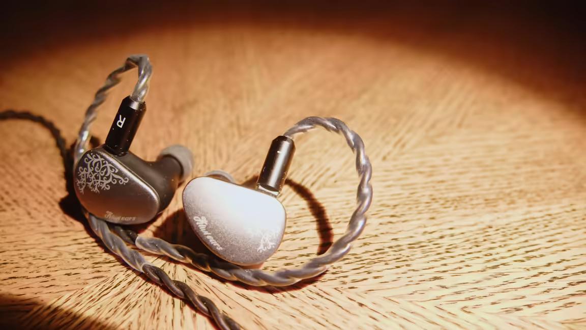The Kiwi Ears Belle's two earpieces on wood - one showing the sunburst metal faceplate, the other the resin body - and its silver-plated copper cable