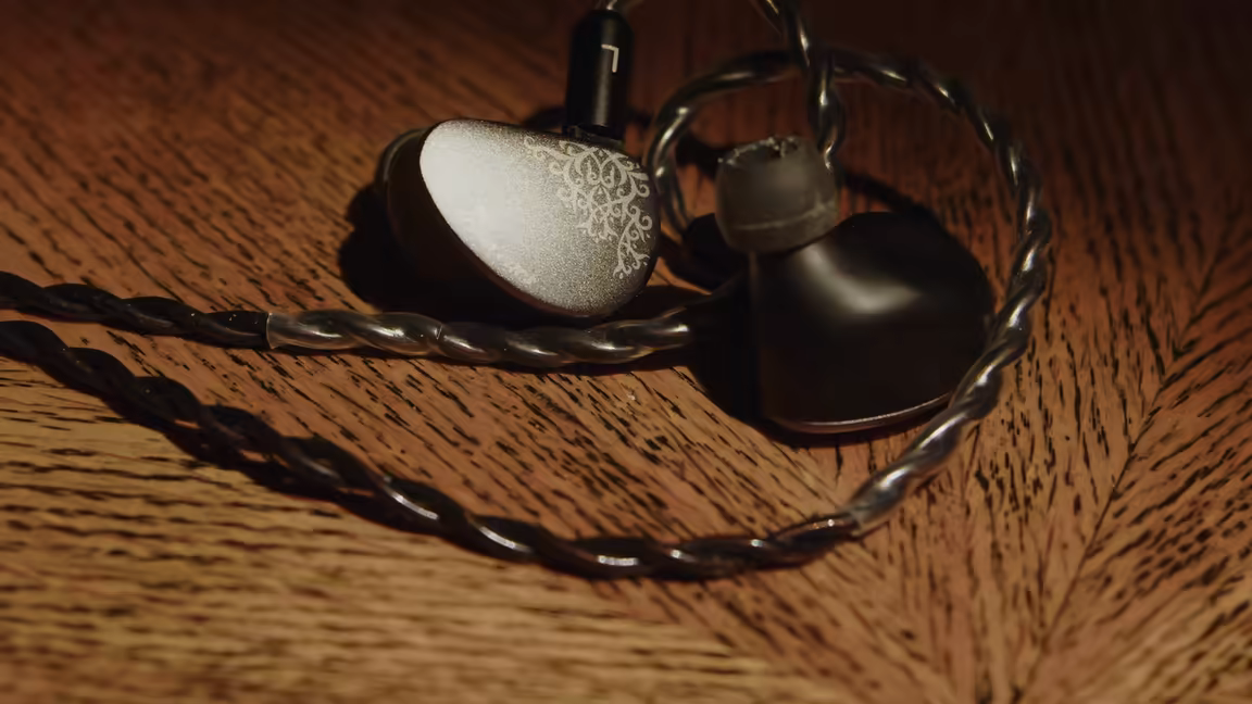 The Kiwi Ears Cadenza II earpieces on wood, one showing its silver filigree faceplate, beside the dark single-crystal copper cable