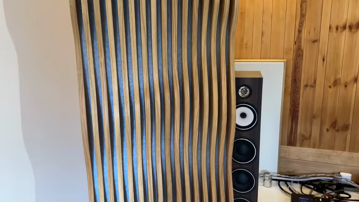 A Parametric Tower standing free in the listening room beside a floor-standing speaker, the dark felt core visible between its oak slats