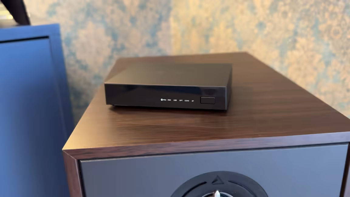 The SMSL PS200 resting on top of a wooden bookshelf speaker, showing how small it is, with its front input-indicator LEDs facing out