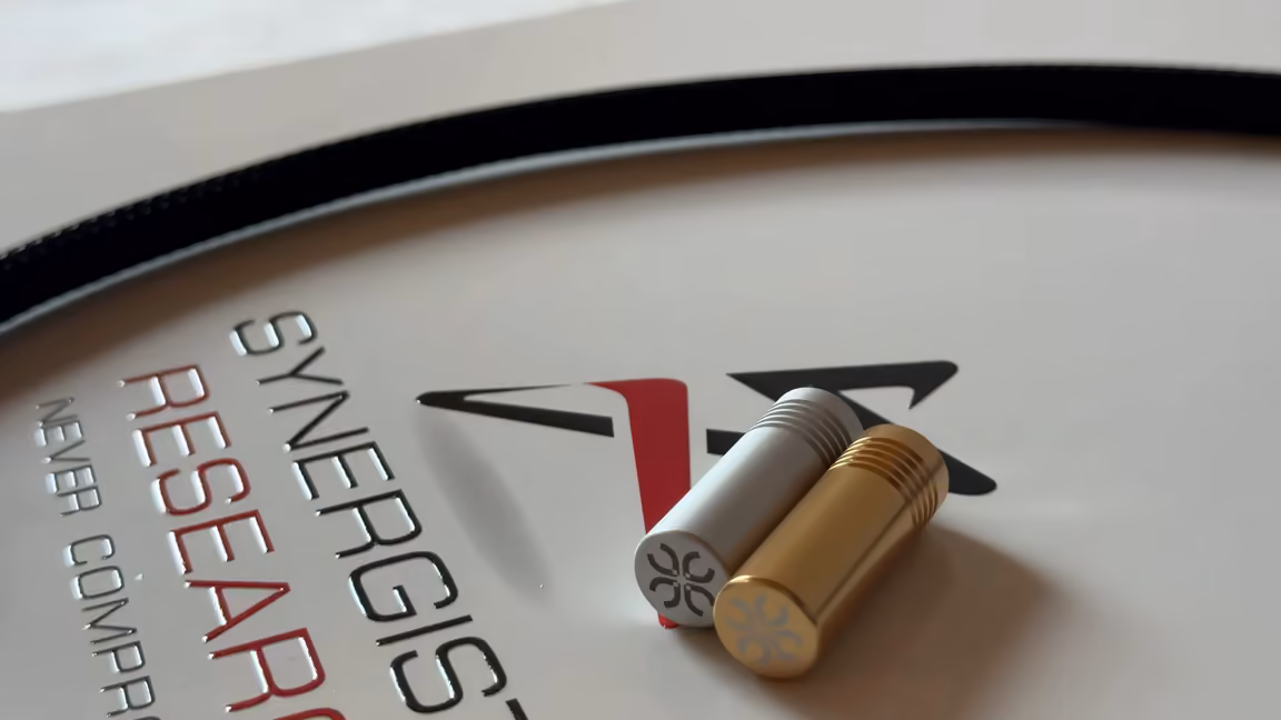 Two Synergistic Research UEF tuning bullets, one silver and one 24-karat gold, machined cylinders with knurled rings and the company's four-petal UEF mark on each flat end cap, resting on the gloss white box beside the coiled power cable