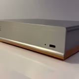The Henry Audio DA 256 seen from the front and side: a low brushed aluminum box with the engraved Henry Audio logo, a solid wood base underneath, and one tiny LED beside a USB-C port on the front plate