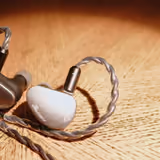 The Kiwi Ears Belle's two earpieces on wood - one showing the sunburst metal faceplate, the other the resin body - and its silver-plated copper cable