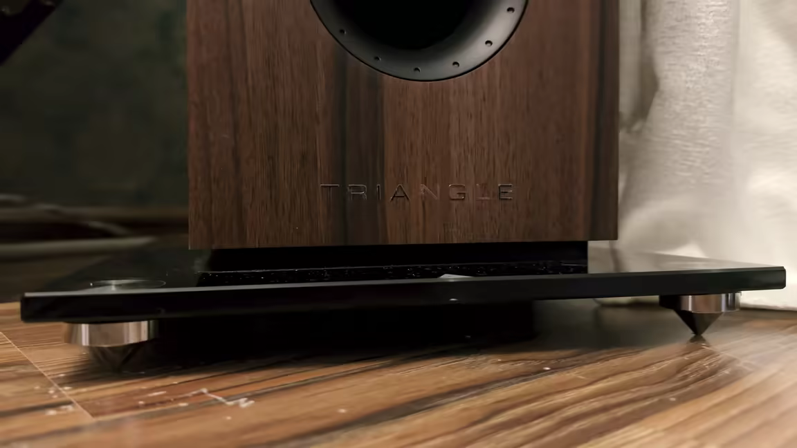 The base of the Australe EZ on its glossy black glass pedestal with chrome spike feet, the Triangle name embossed in the chestnut veneer above